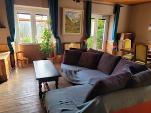 Une pièce chaleureuse avec un plancher en bois, un canapé, une table et une chaise. Il y a des fenêtres avec des rideaux, un tableau au mur et une plante.