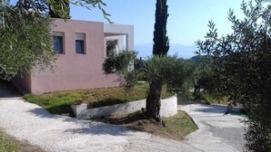 A modern building with a pink facade, a gravel driveway, and surrounding greenery. - Wohlfühloase Inmitten Alter Oliven- und Zypressenbäumen (Karousades)