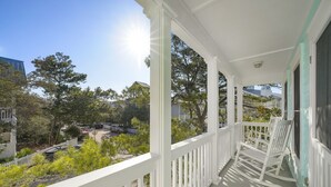 A covered porch with white railings and a rocking chair, overlooking a residential area with trees and houses.