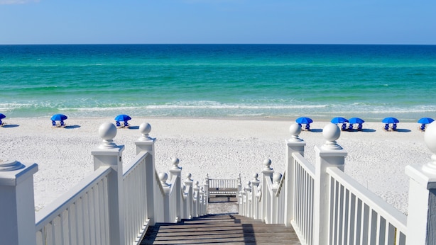 A wooden staircase leading to a beach with white railings and blue umbrellas.