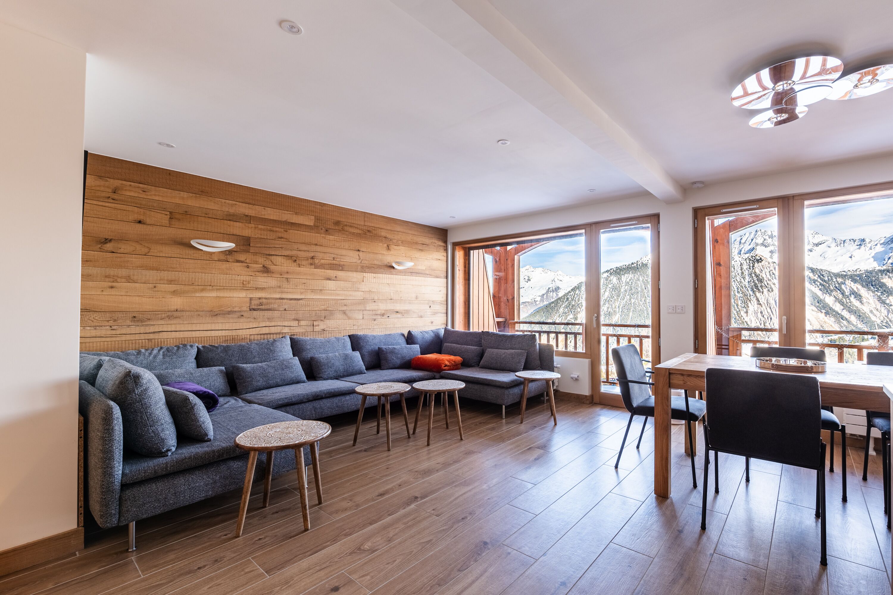 A modern living room with a wooden wall, a grey sofa, small round tables, and a dining area with chairs.