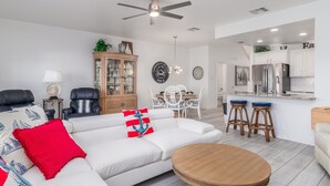 Un salon moderne avec un canapé blanc, des coussins rouges et bleus, une table basse en bois et un coin repas avec des chaises blanches.
