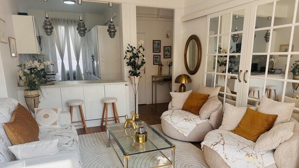 A modern living room with a glass coffee table, beige sofas with gold accents, and a kitchen in the background.