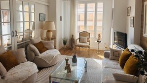 A spacious living room with a sofa, armchair, and a glass coffee table. There is a television, a lamp, and a statue on a side table.