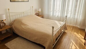A neatly made bed with a beige cover, a bedside table with a lamp, a wooden floor, and a window with sheer curtains.