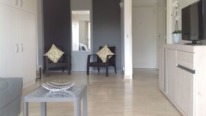 A modern living room with a grey sofa, a small table, two armchairs with patterned cushions, a TV on a cabinet, and a clock on the wall. - Spacious studio in the center of Cannes, nice terrace, easy walk to the Croisette and Palais - 1923 (Cannes)