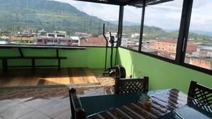 A balcony with a wooden floor, a table with a patterned tablecloth, and a view of a cityscape with buildings and hills. - macas center (Macas)