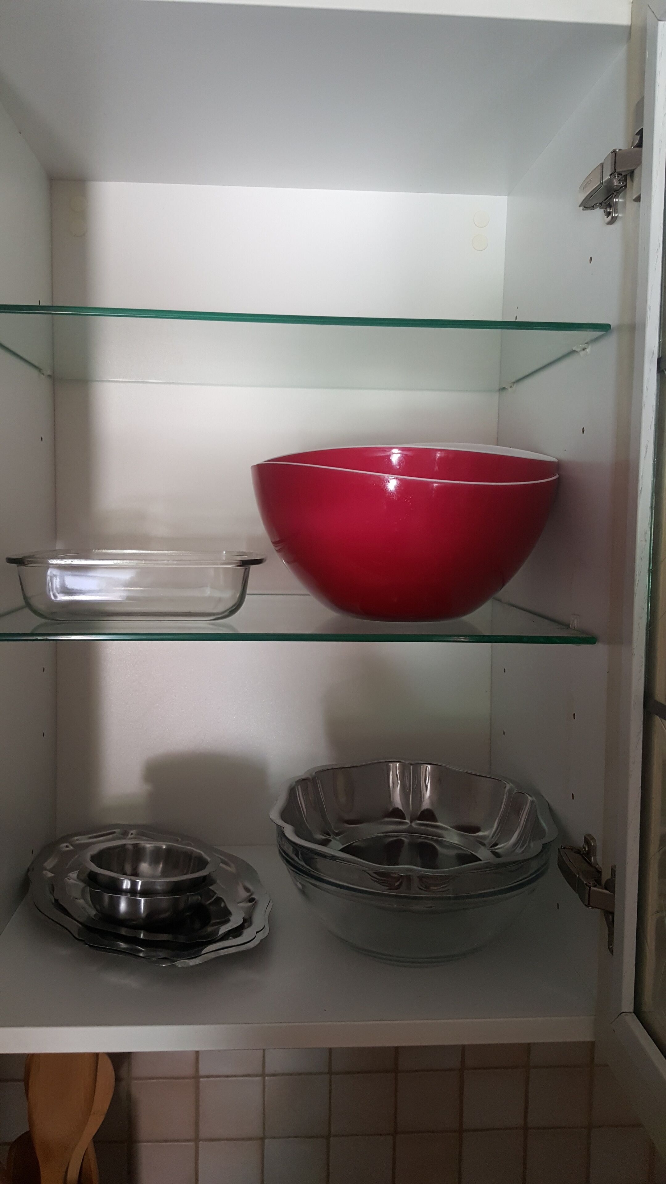Een keuken met een rode kom op het bovenste schap, een glazen kom op het middelste schap, en een metalen kom op het onderste schap.