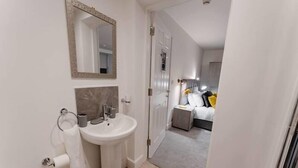 Une salle de bain moderne avec un lavabo, un miroir et des toilettes. Une chambre à coucher avec un lit et une table de chevet est visible à travers une porte ouverte.