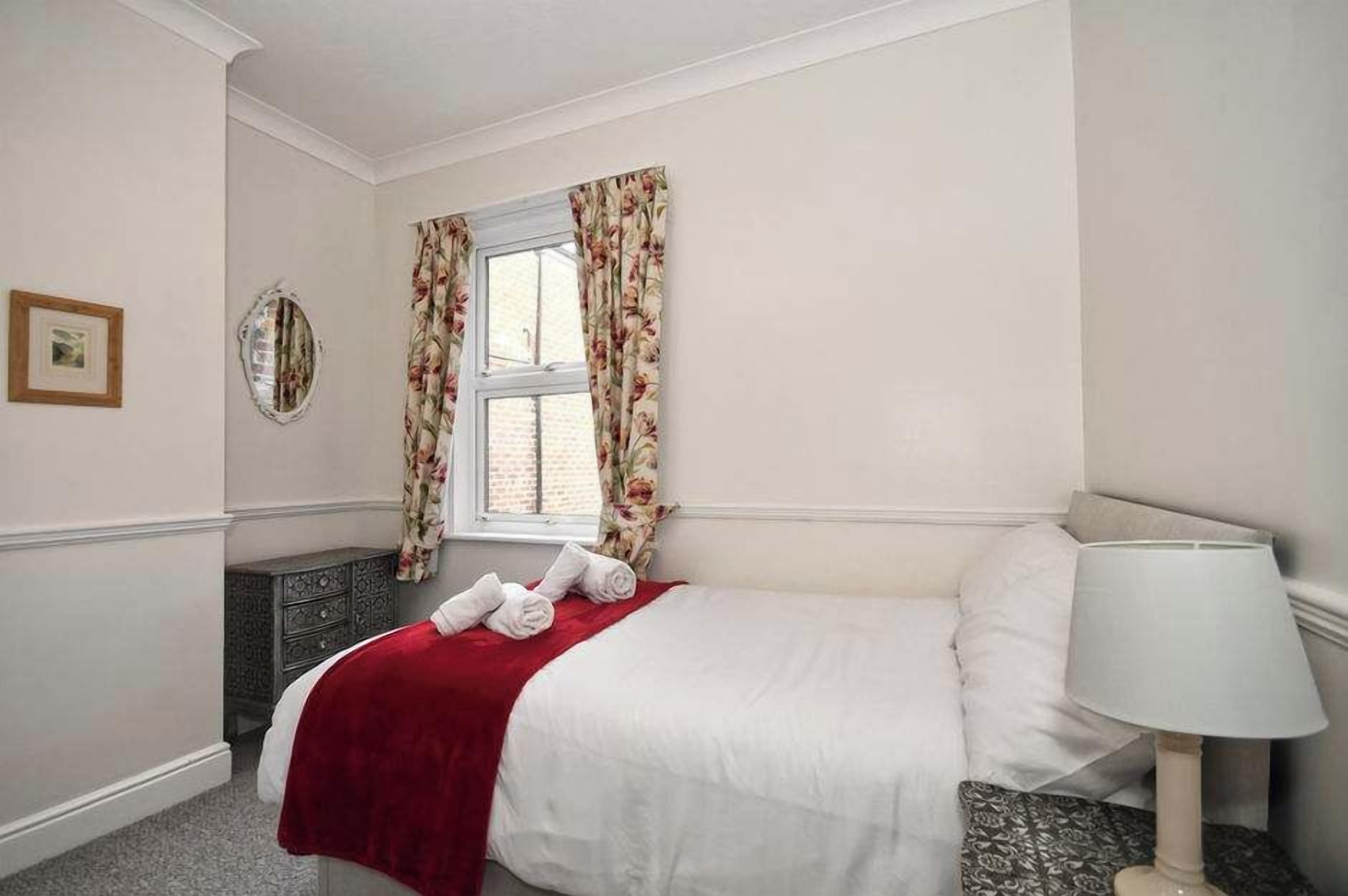 A hotel room with a bed, a bedside lamp, a dresser, a window with curtains, and a mirror on the wall.