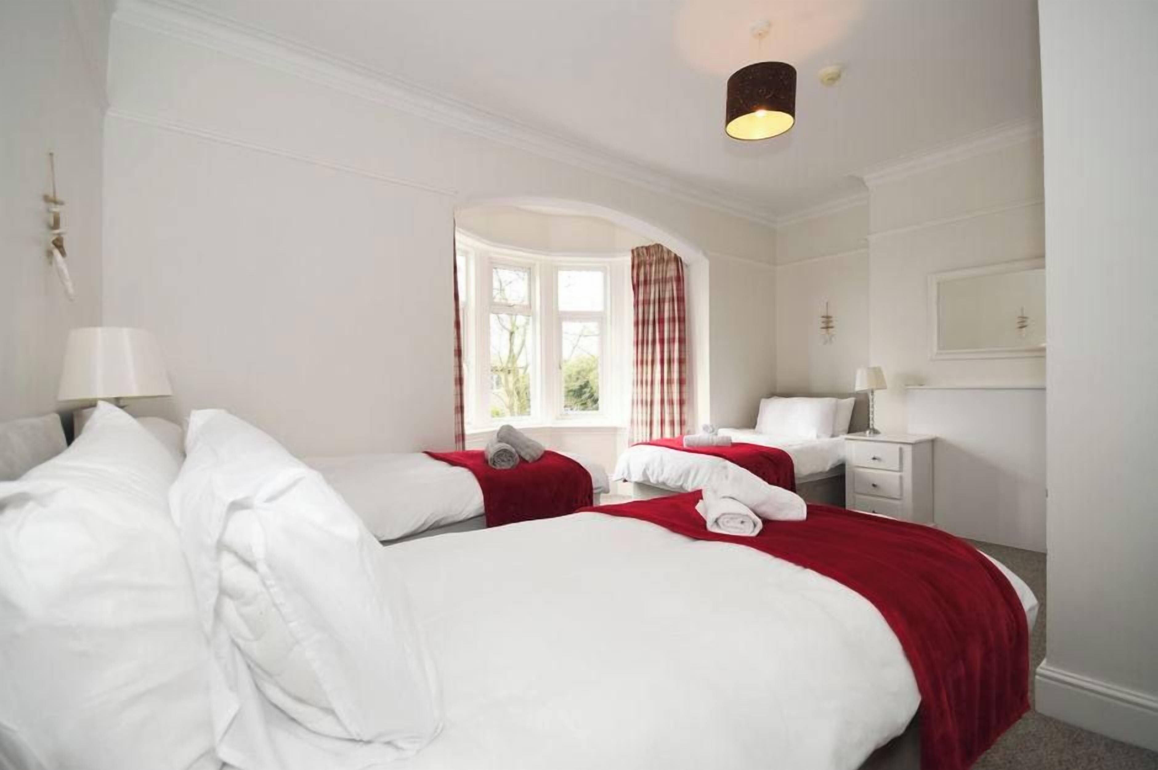 A hotel room with two beds, a window with curtains, a bedside table with a lamp, and a mirror on the wall.