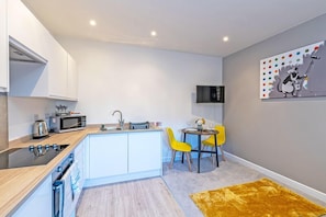 A compact kitchen with a microwave, kettle, and sink. There is a small dining area with a table and two yellow chairs. A painting of a cat on a roller is on the wall.