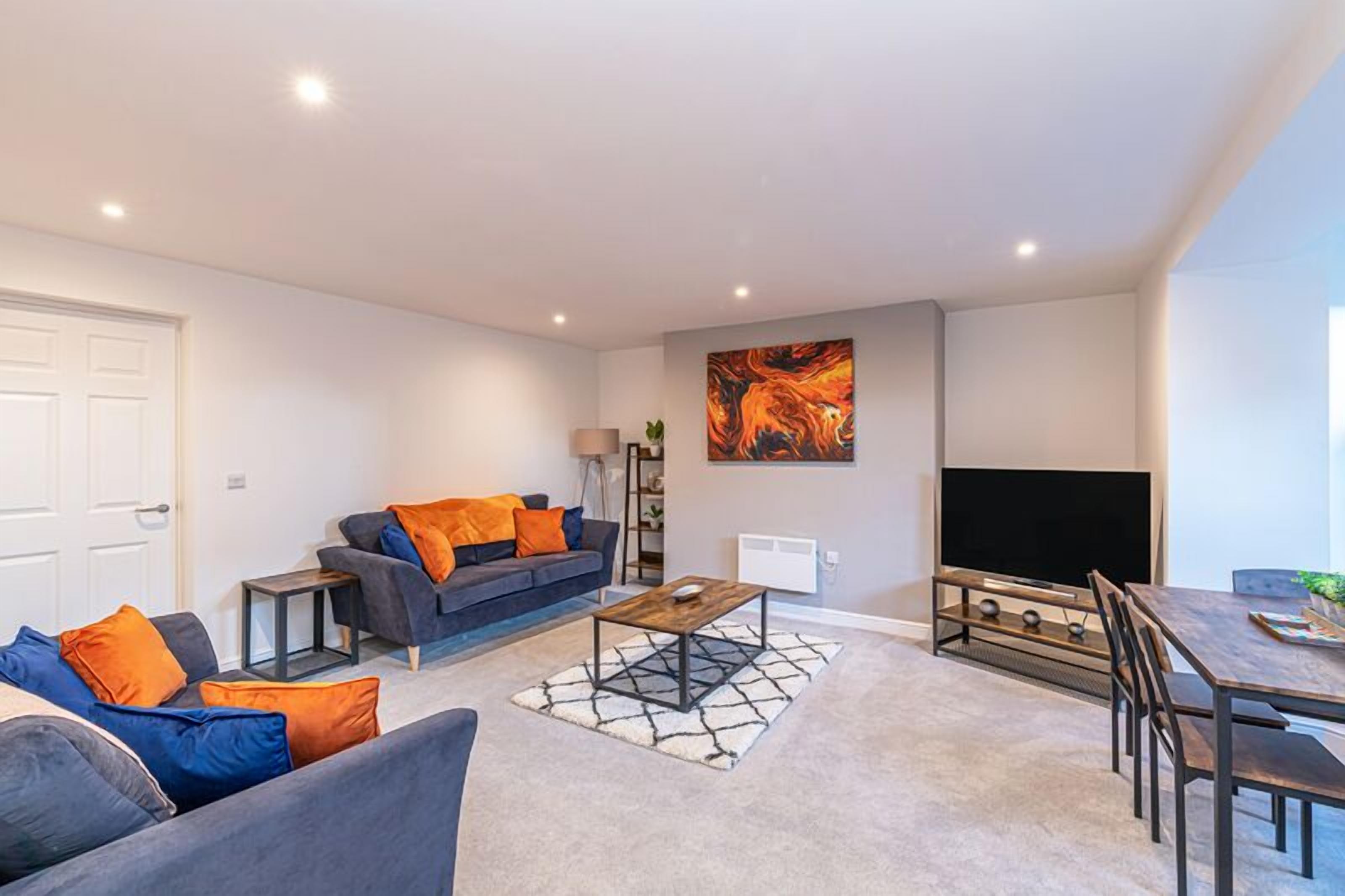 A modern living room with a grey sofa, orange cushions, a coffee table, a flat-screen TV, and a dining area with a wooden table and chairs.