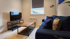 Un salon avec un canapé foncé, une table basse en bois, un téléviseur à écran plat et une fenêtre équipée d’un store.
