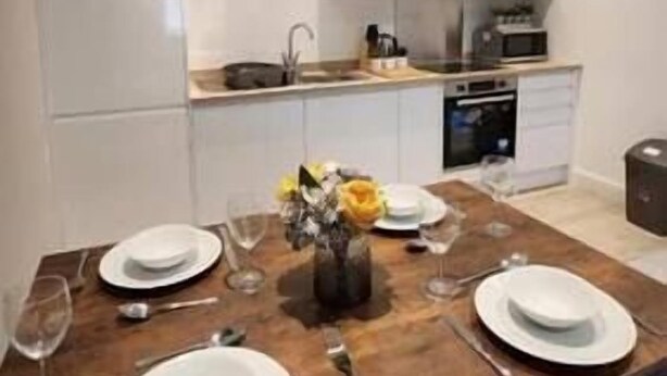 Une cuisine moderne avec des meubles blancs, une table à manger en bois dressée pour quatre personnes et un vase rempli de fleurs jaunes.