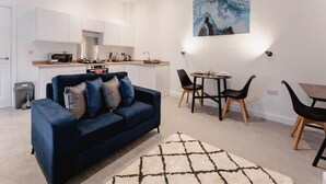 En moderne stue med en mørkeblå sofa, et spiseområde med et lille bord og stole, et køkken med hvide skabe, og et billede på væggen.