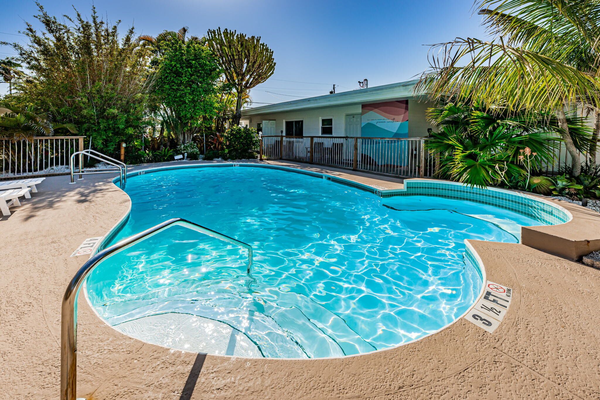 A swimming pool with a curved design, surrounded by a sandy deck and a fence.