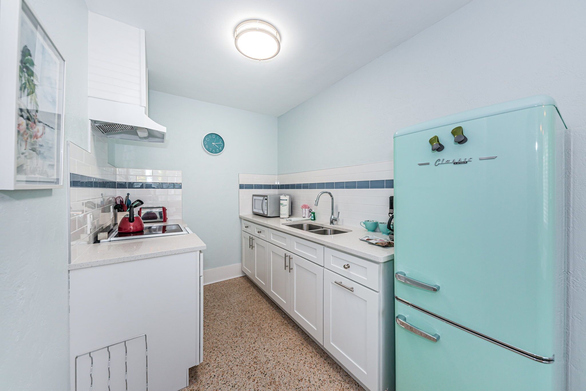 Een compacte keuken met witte kasten, een blauwgroene koelkast, een magnetron, een broodrooster en een spoelbak.
