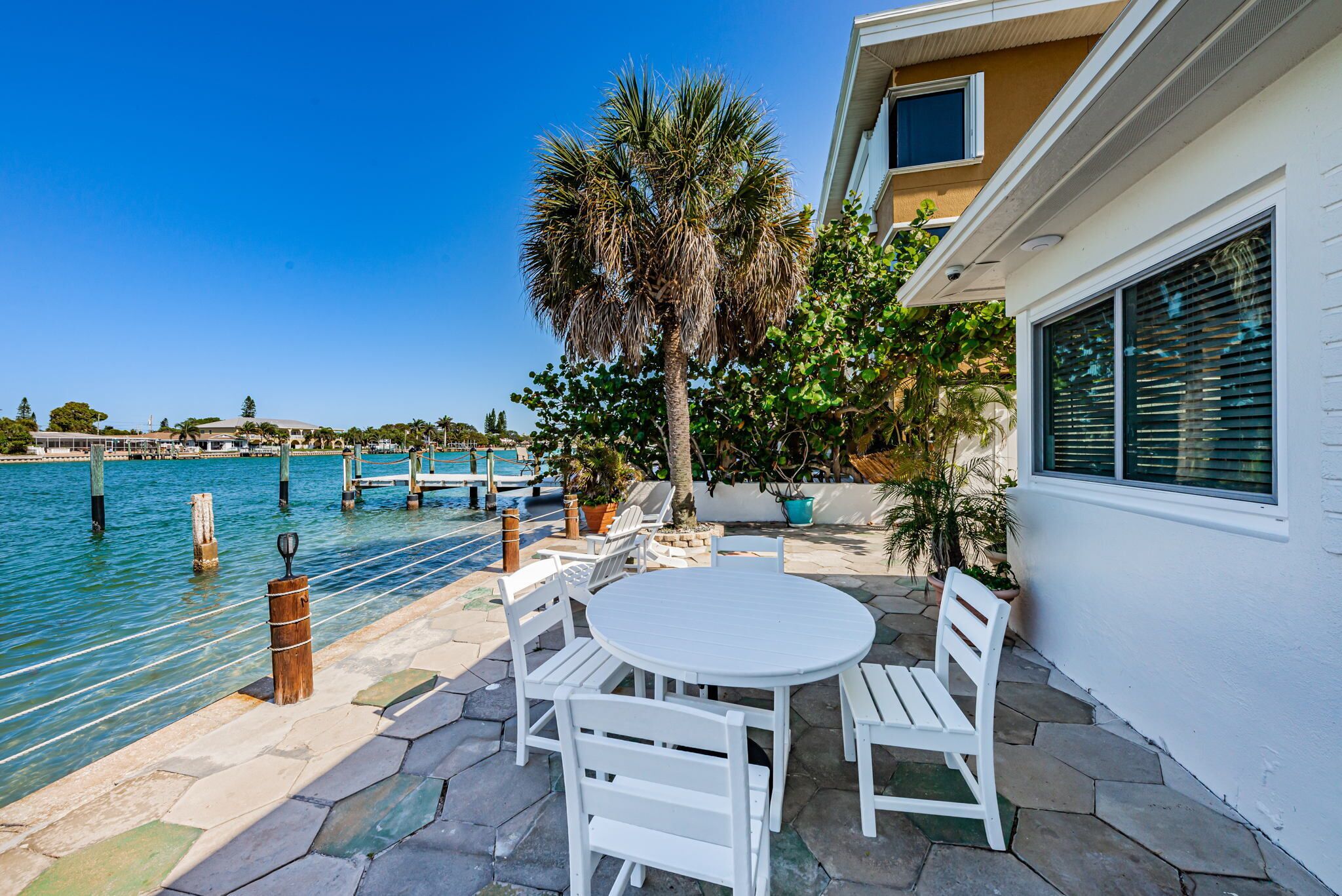A patio with a round table and white chairs, overlooking a body of water with a dock and palm trees.
