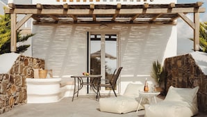 Un patio soleggiato con un muro in pietra, un bagno esterno bianco, un pergolato in legno, un tavolo con sedie e una zona salotto con cuscini.