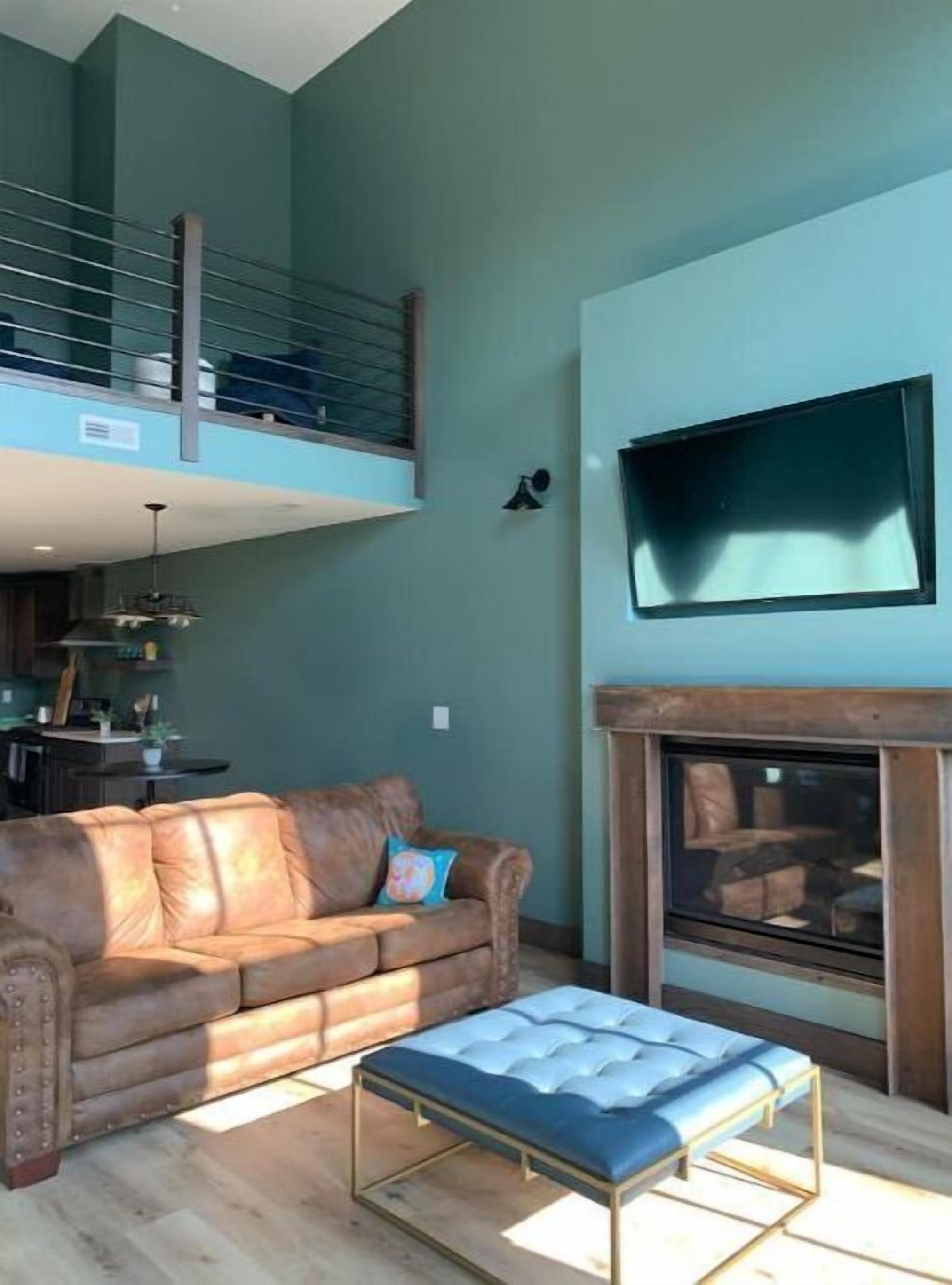 A spacious living room with a high ceiling, a leather sofa, a blue ottoman, a fireplace, and a television mounted on the wall.