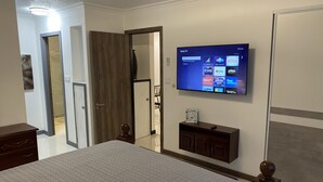 A hotel room with a large bed, a TV mounted on the wall displaying streaming channels, a wooden cabinet below the TV, and a door leading to another room.