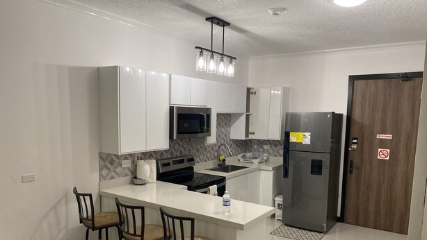 A modern kitchen with white cabinets, a stainless steel refrigerator, a microwave, and a bar area with stools.