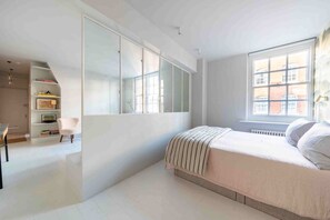 A bedroom with a large window, a bed with a striped blanket, a chair, and a bookshelf. - Open plan Chelsea apt for 2 nr King’s Road (London)