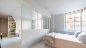 A bedroom with a large window, a bed, a chair, and a built-in shelf unit. - Open plan Chelsea apt for 2 nr King’s Road (London)
