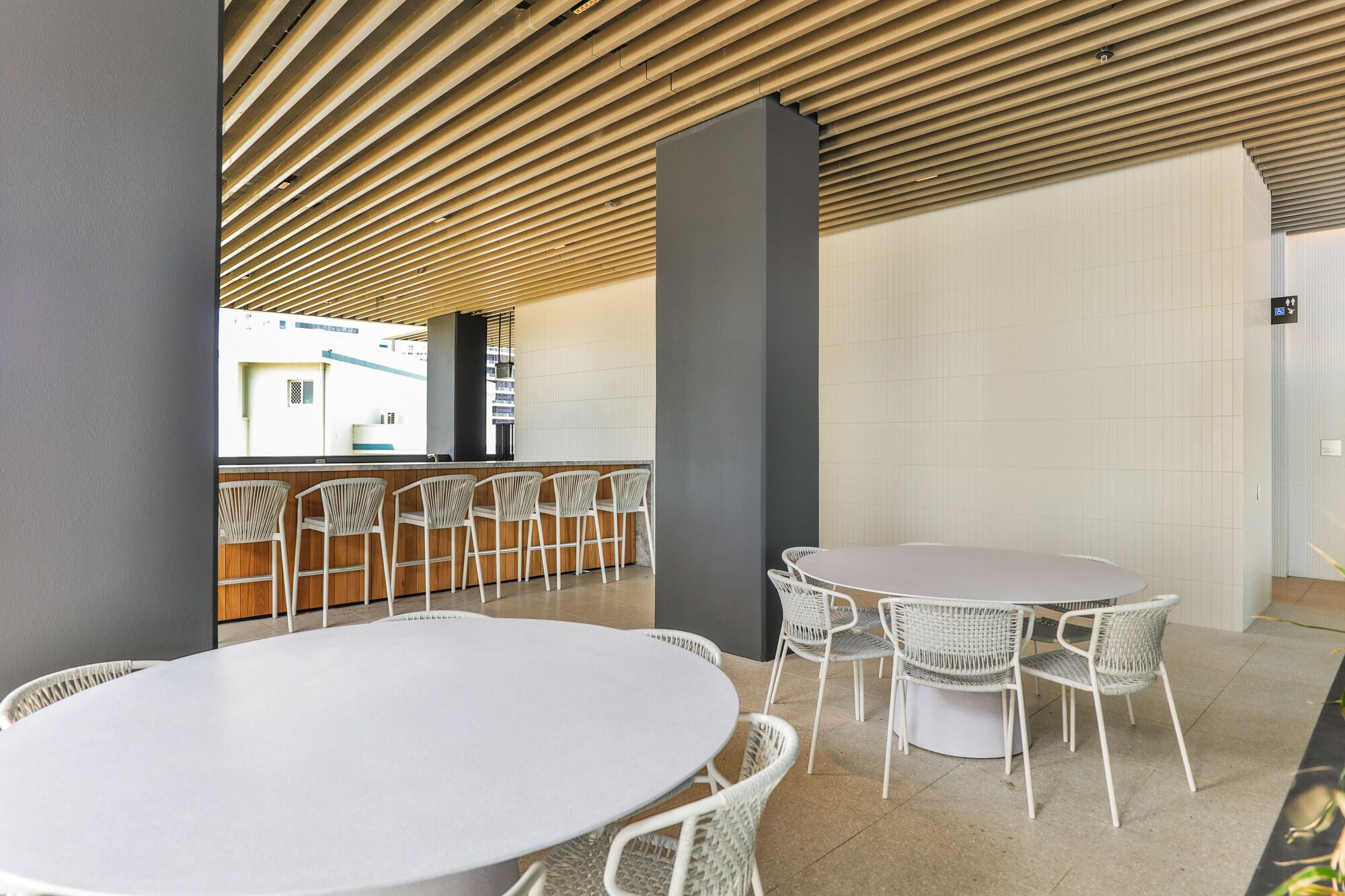 A modern indoor dining area with round tables and wicker chairs, a wooden slatted ceiling, and a view of the outside.