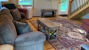 A cozy living room with a gray couch, wooden coffee table, and a large rug. A flat-screen TV is mounted on the wall, and a staircase with a wooden railing is visible in the background.