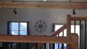A room with a sloped ceiling, a ceiling fan, two windows with curtains, a compass wall decoration, and a wooden beam structure.