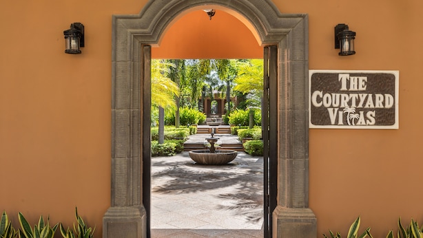 Une entrée surmontée d’une arche qui mène à une cour intérieure agrémentée d’une fontaine et de verdure.