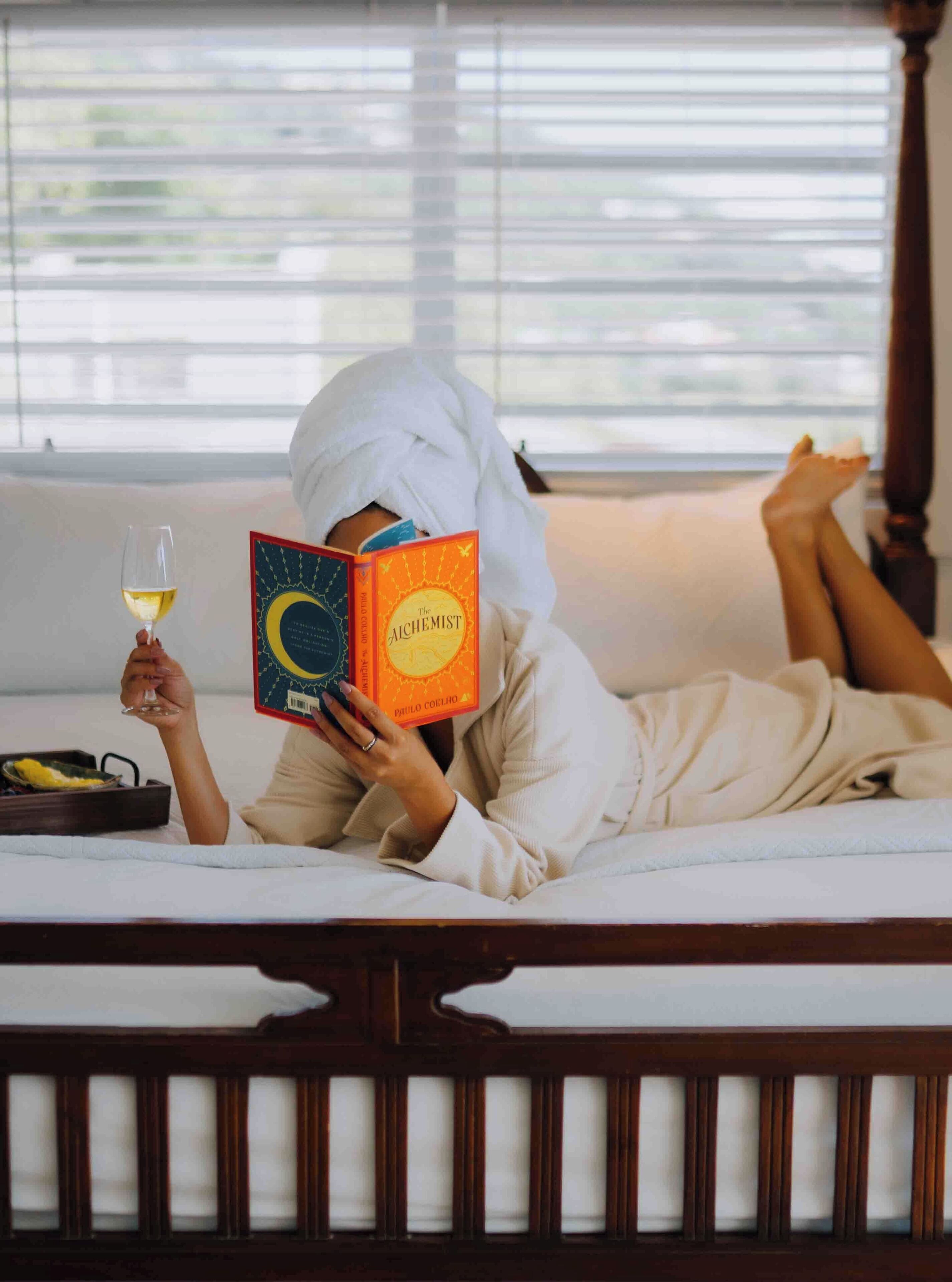Una persona recostada en una cama leyendo un libro titulado 'El Alquimista' de Paulo Coelho, con una copa de vino cerca.