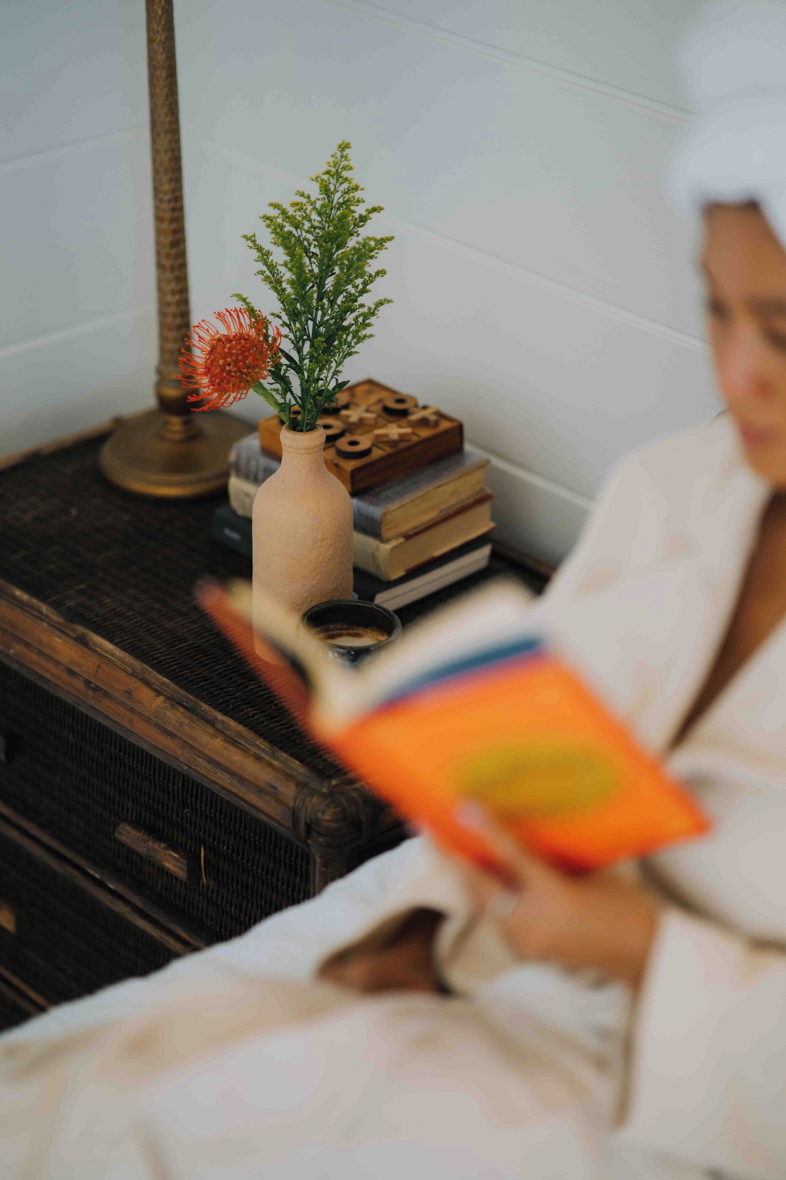 Een persoon leest een boek in een kamer met een rieten stoel, een klein tafeltje met een vaas en boeken, en een lamp.
