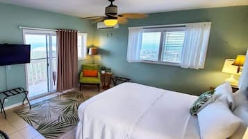 A hotel room with a bed, a ceiling fan, a TV, a window with curtains, a chair, and a small table.