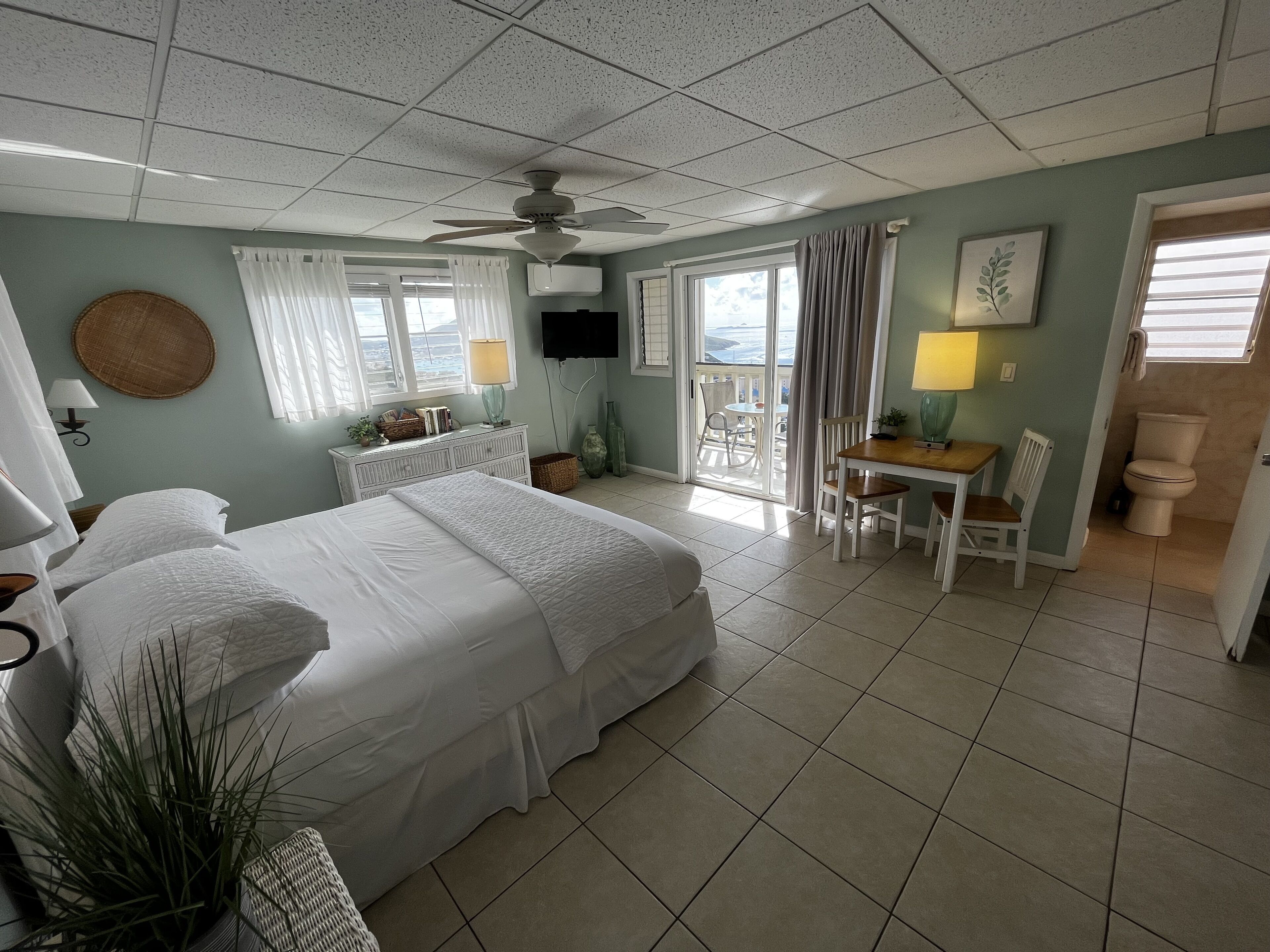 A hotel room with a bed, a desk, a chair, a ceiling fan, and a view of the outdoors.