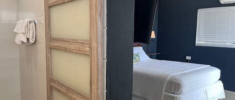 A bedroom with a bed, a window with blinds, a towel rack, and a wooden-framed door.