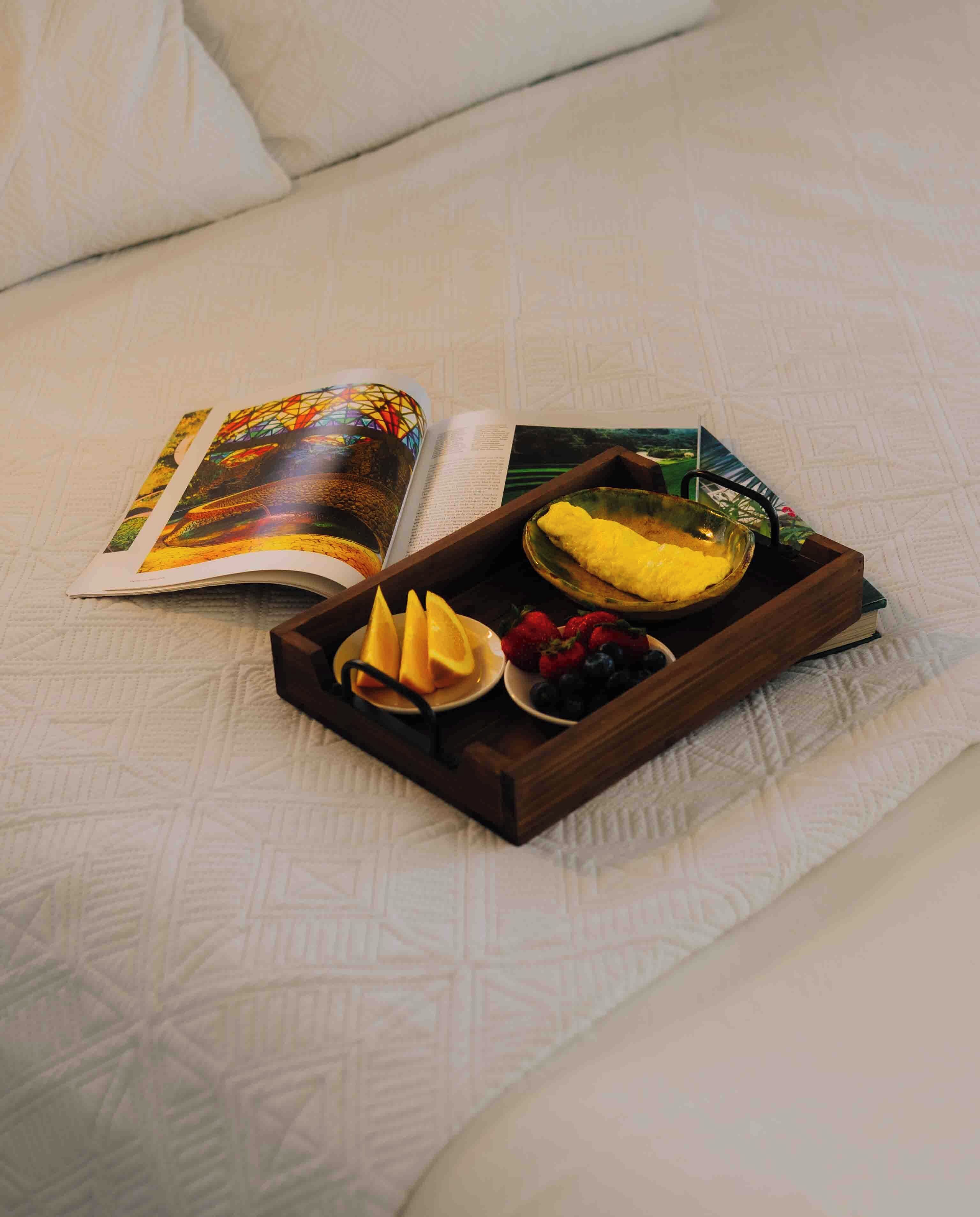 Ein Bett mit einer gesteppten Musterung, ein Tablett mit Früchten und einem Buch, sowie eine Zeitschrift mit farbigem Einband.