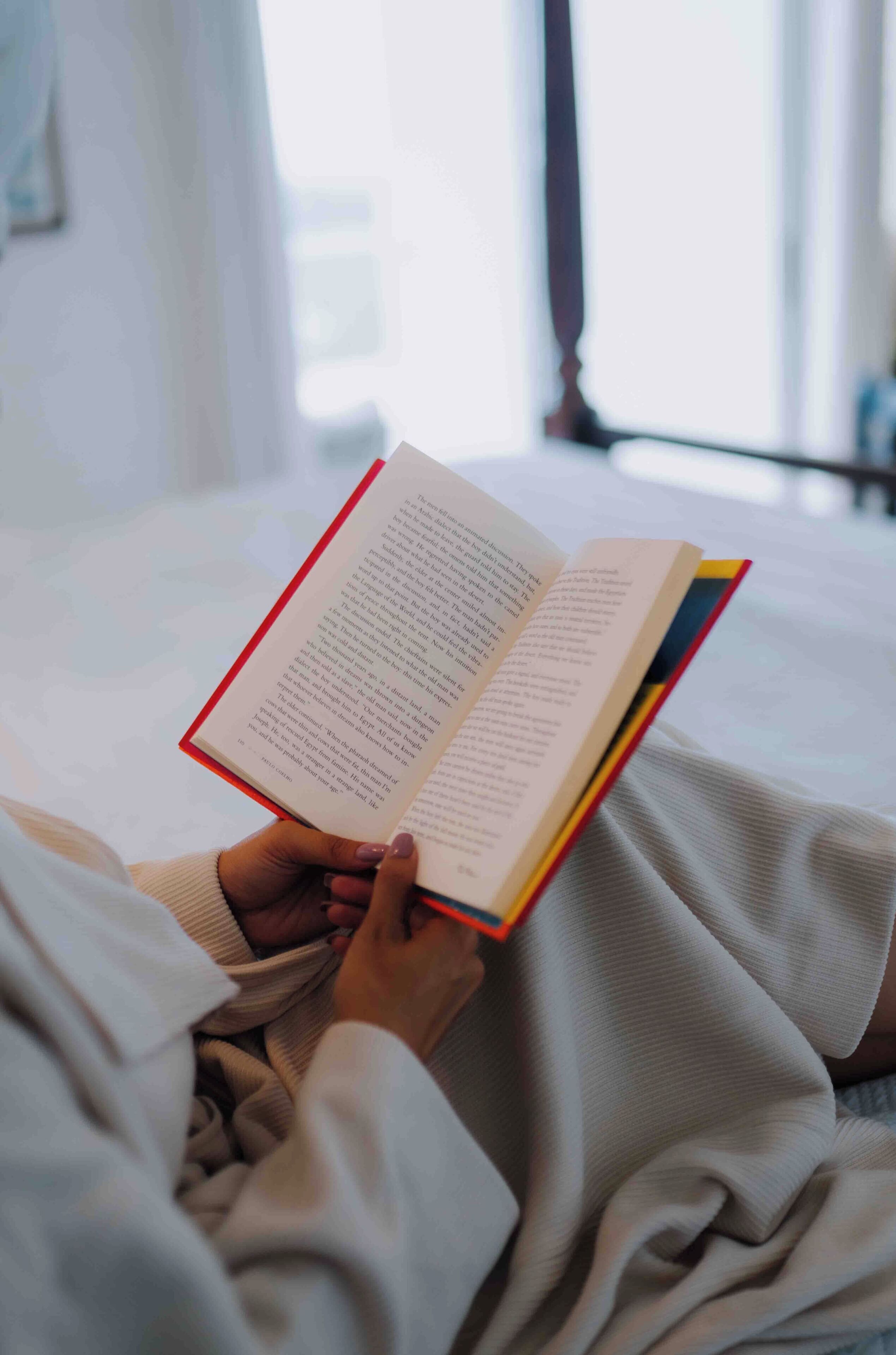 A person reading a book in a room with a bed and a window.