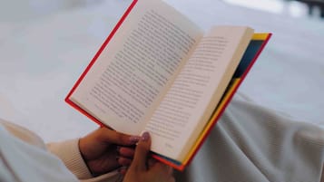 Una persona leyendo un libro en una habitación con cama y ventana.
