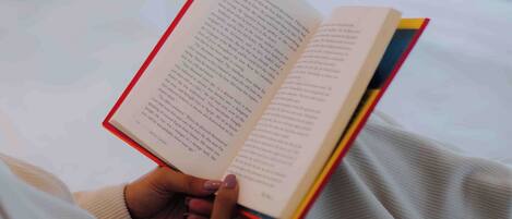 A person reading a book in a room with a bed and a window.