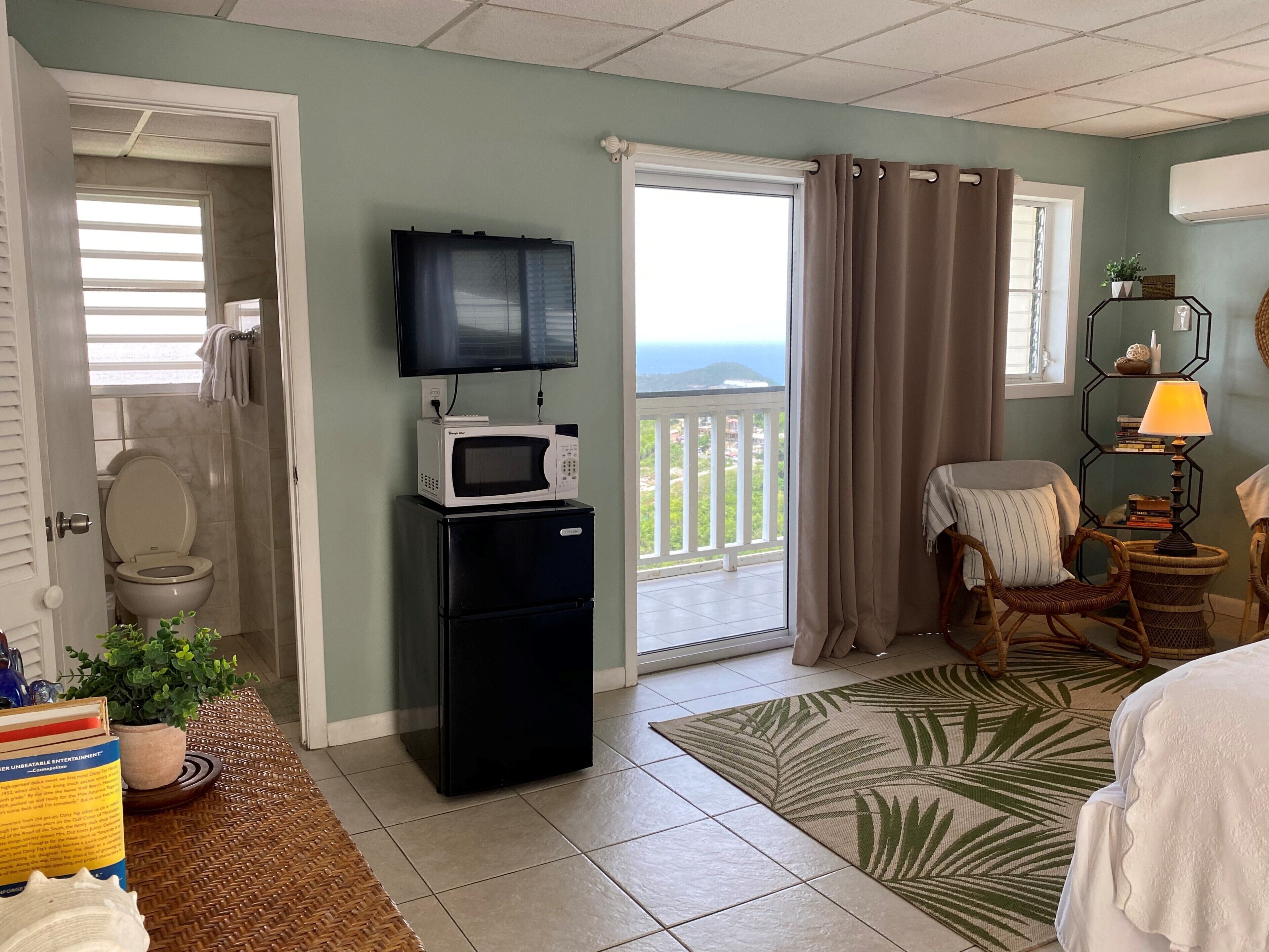A hotel room with a balcony, a television, a microwave, a refrigerator, a wicker chair, a small table with a lamp, and a view of the ocean.
