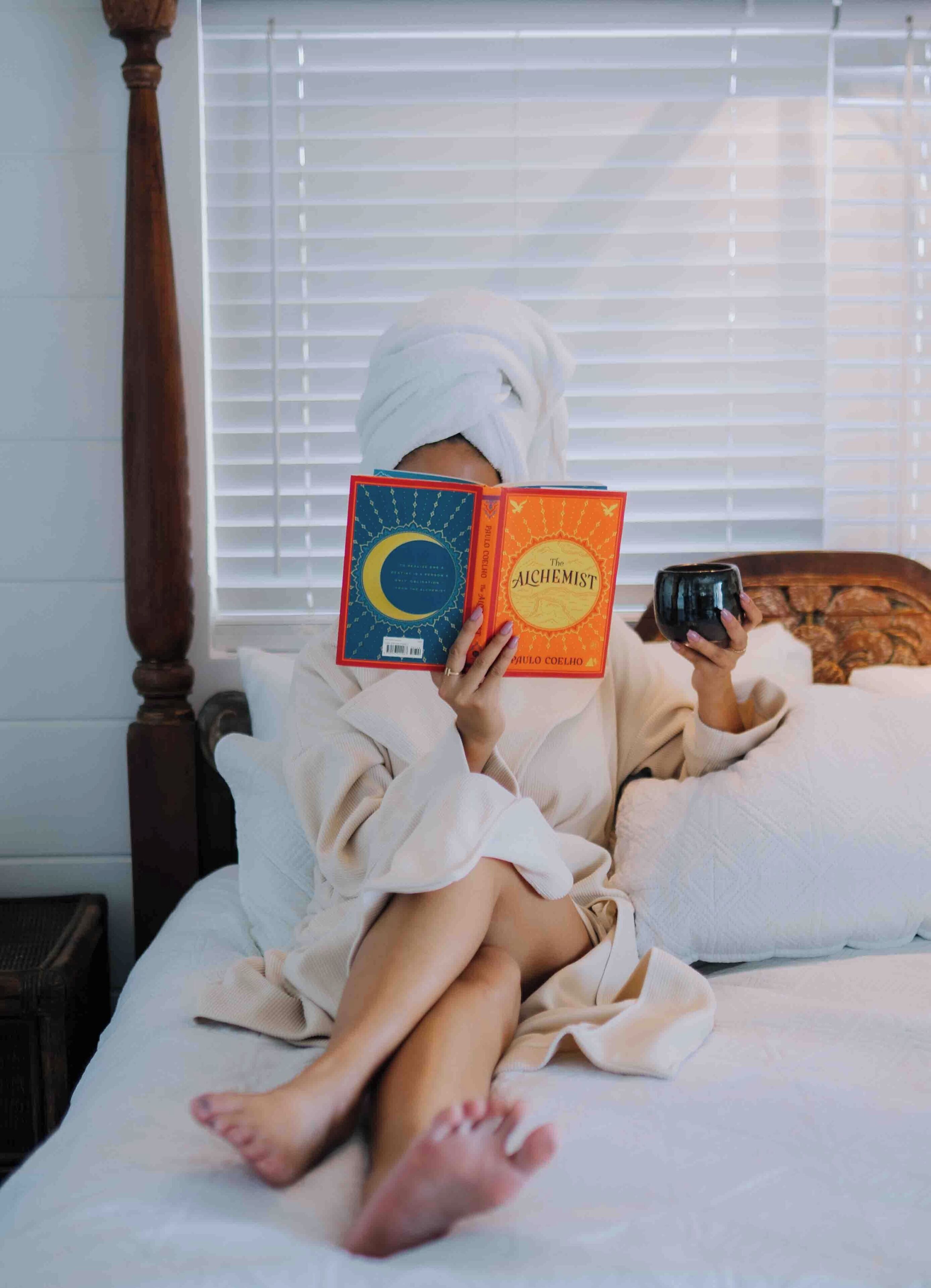 Una persona leyendo un libro en una cama con cabecera de madera, una ventana con persianas y una mesita de noche.