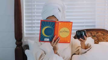 Una persona leyendo un libro en una cama con cabecera de madera, una ventana con persianas y una mesita de noche.