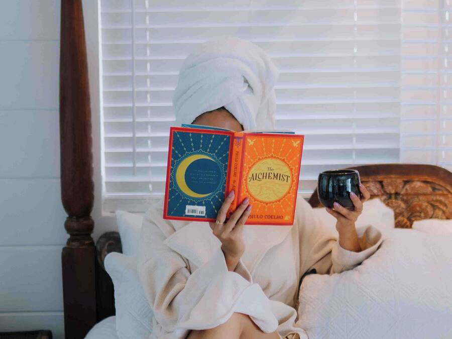 Una persona leyendo un libro en una cama con cabecera de madera, una ventana con persianas y una mesita de noche.