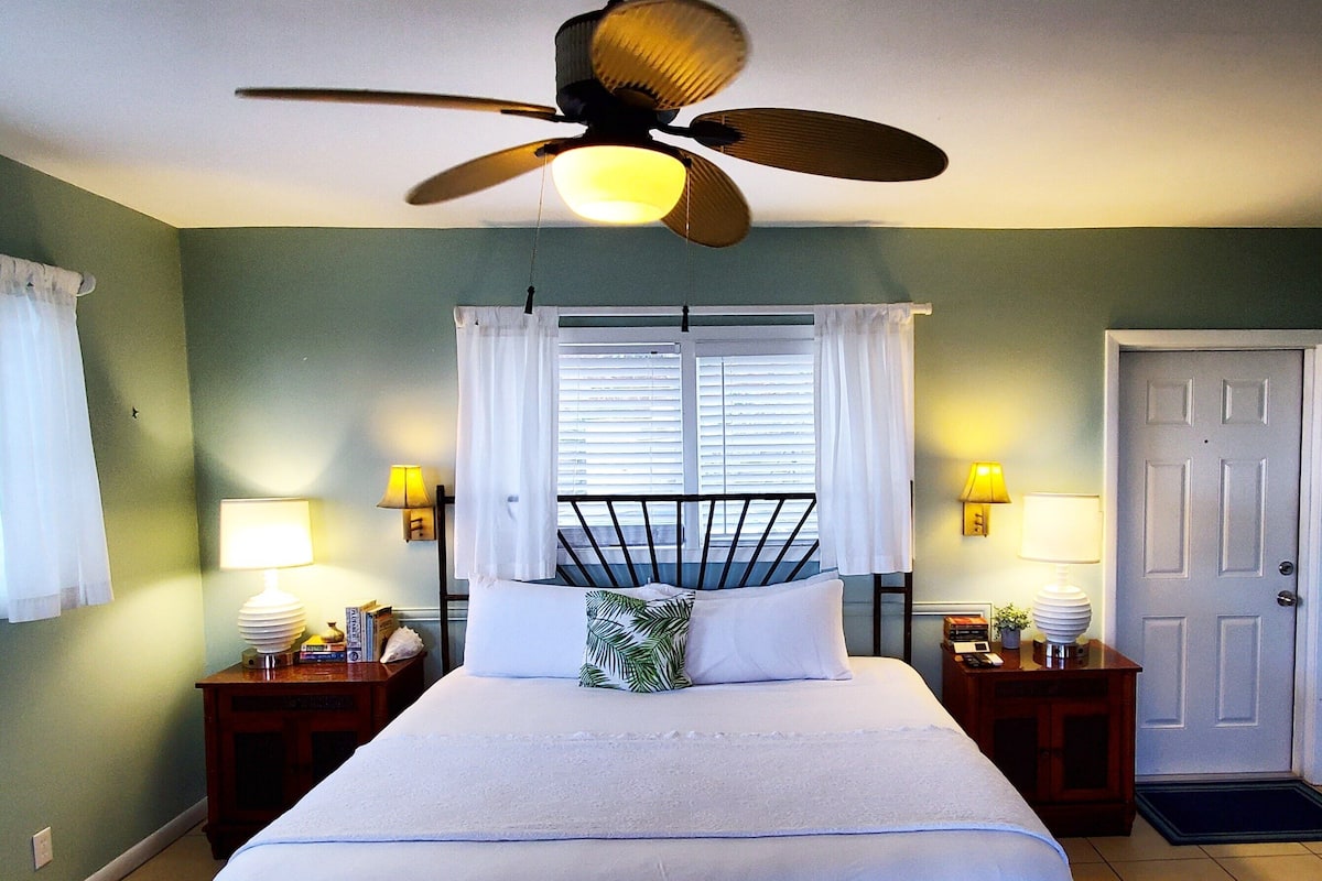 A bedroom with a bed, two bedside tables with lamps, a ceiling fan, a window with blinds, and a door.