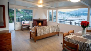 A room with a fireplace, a dining table with a vase of flowers, and a view of a marina. - White Heaven Marina View. Studio Near the Lakki Port. (Lakki)