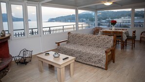 A spacious room with a sofa, dining table, and chairs. Large windows offer a view of a body of water and distant hills. - White Heaven Marina View. Studio Near the Lakki Port. (Lakki)