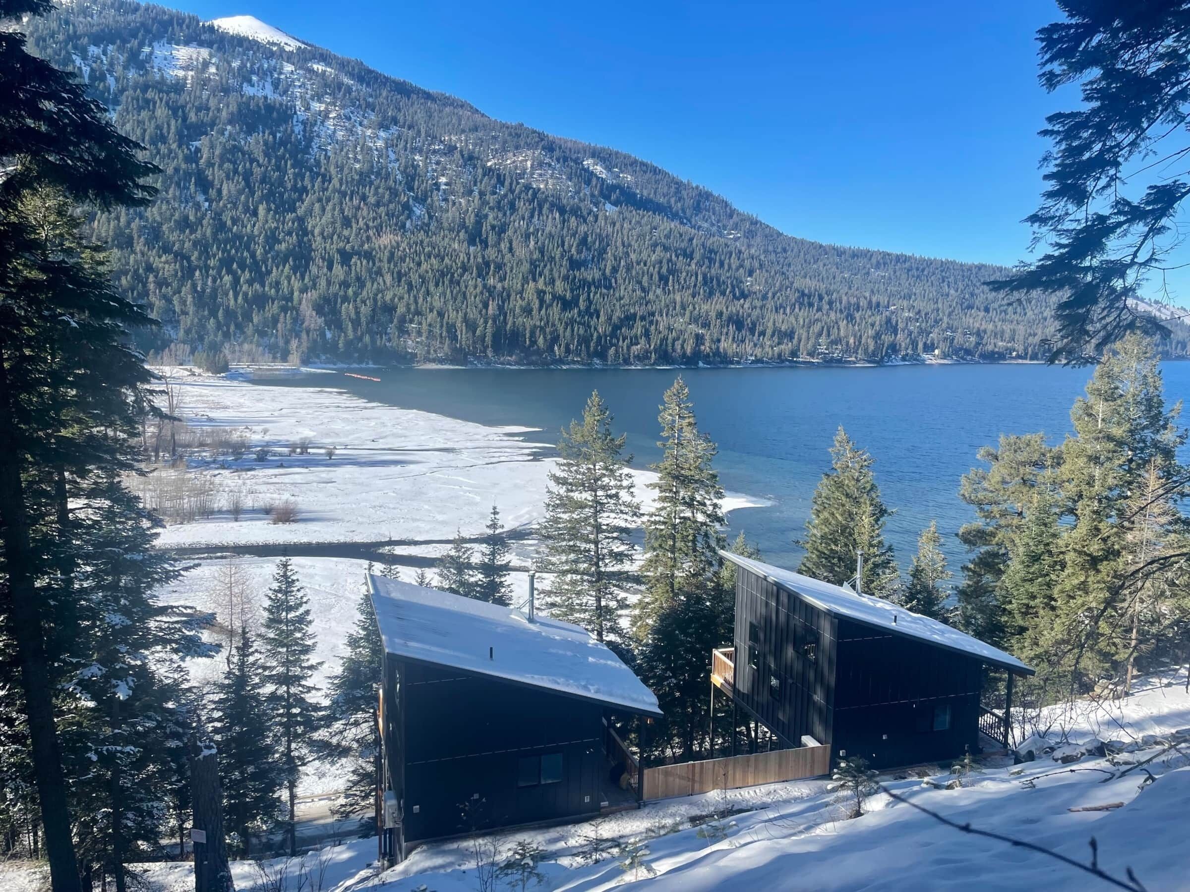 Une cabane dont le toit est recouvert de neige, entourée de pins, et qui offre une vue sur un lac et des montagnes.